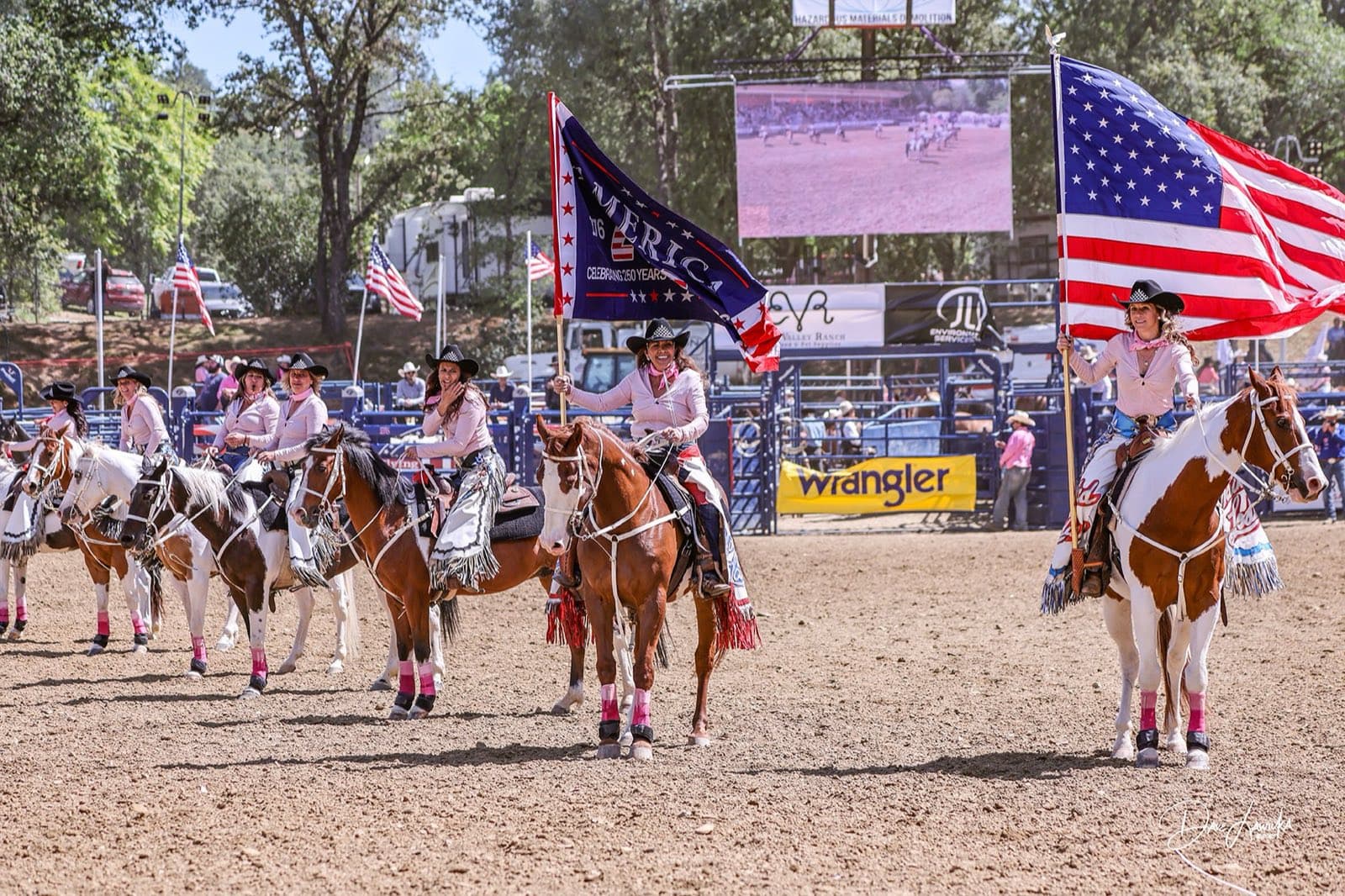 Rodeo queens in pink shirts on horseback carrying the American flag in the grand entry at McCann Stadium
